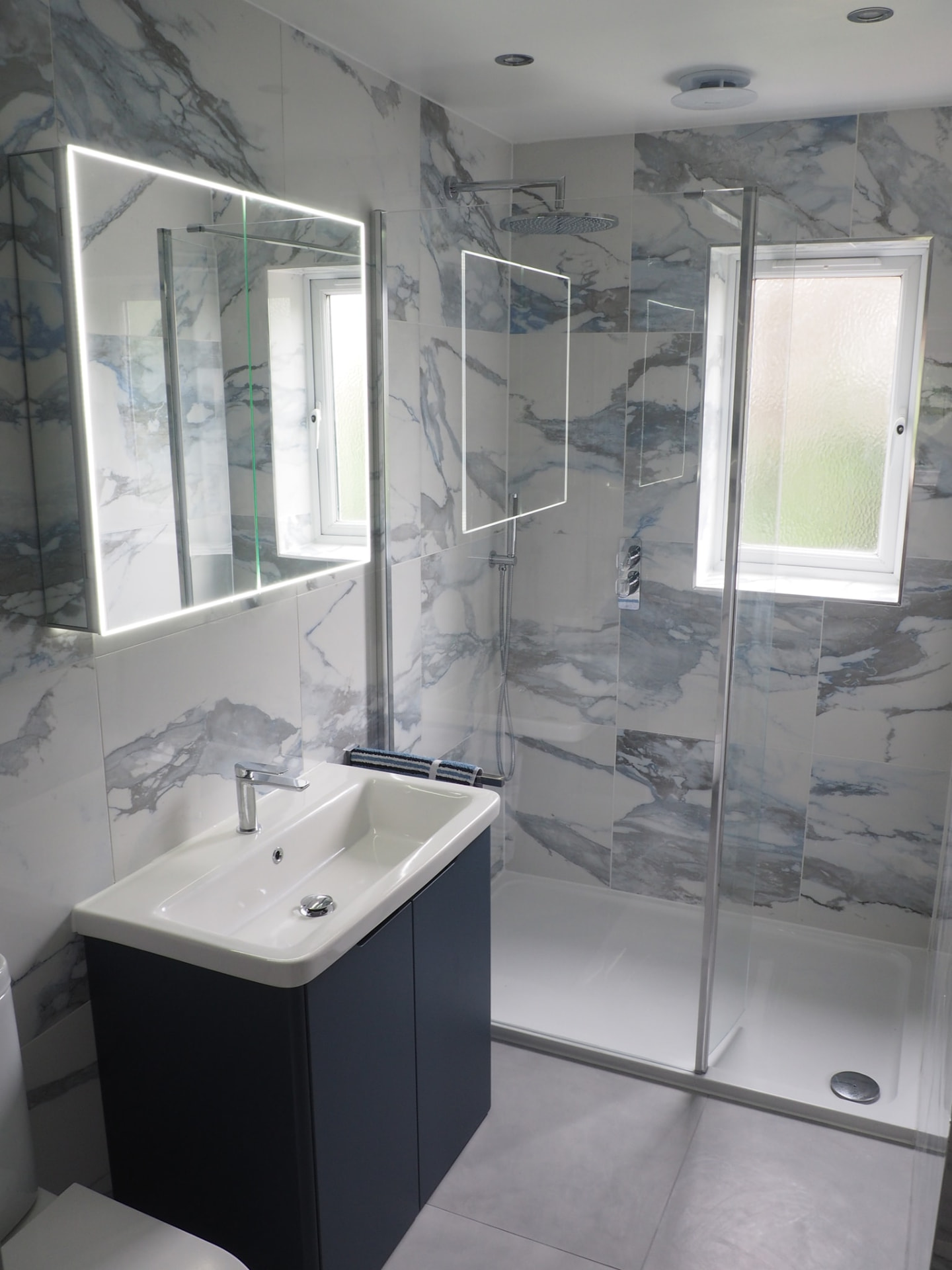 Bathroom with blue-veined marble tiling, LED mirror cabinet, navy vanity unit and walk-in rainfall shower